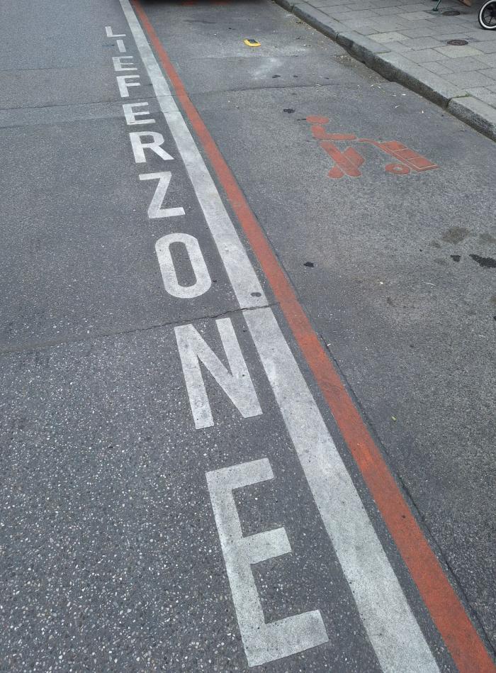Das Foto zeigt den Schriftzug Lieferzone in weiß von oben nach unten neben der in rot markierten Lieferzone, die einen Parkplatz umrandet.