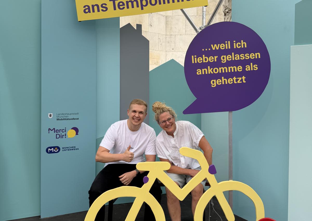 Ein Mann und eine Frau sitzen im Pop-up-Fotostudio von Merci Dir hinter einem Holzfahrrad. Ihr Statement: "Bitte haltet euch ans Tempolimit – weil ich lieber gelassen ankommen, als gehetzt"