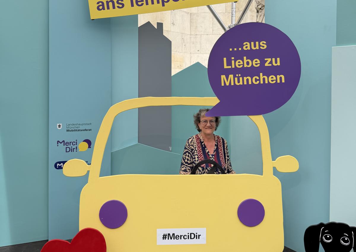 Eine Frau sitzt im Pop-up-Fotostudio von Merci Dir in einem Holzauto. Ihr Statement: "Ich halte mich ans Tempolimit – aus Liebe zu München"