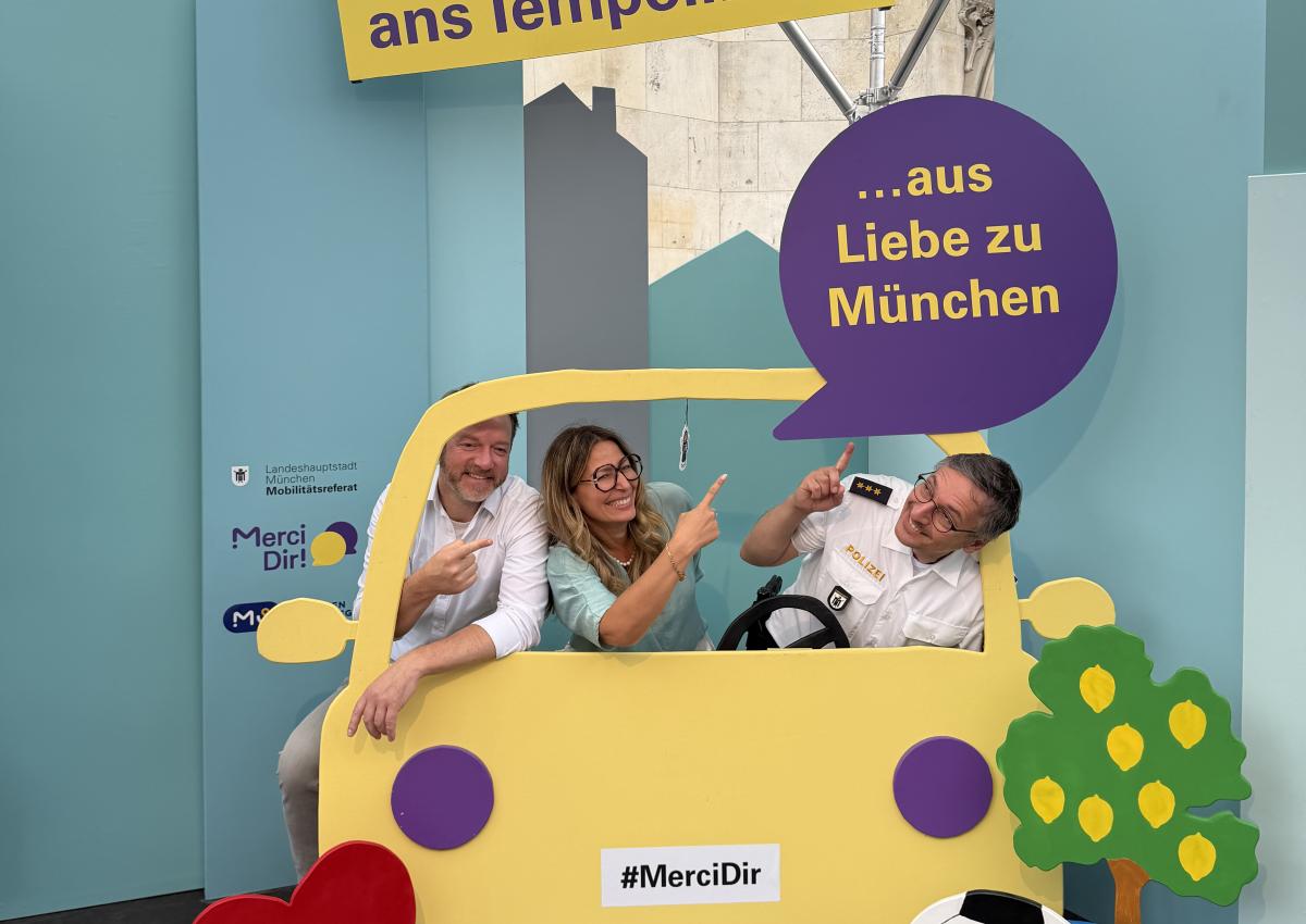 Zwei Männer und eine Frau sitzen im Pop-up-Fotostudio von Merci Dir in einem Holzauto. Ihr Statement: "Ich halte mich ans Tempolimit – aus Liebe zu München"
