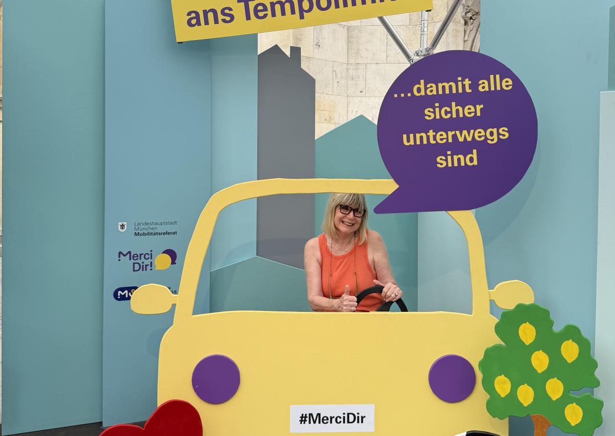 Eine Frau sitzt im Pop-up-Fotostudio von Merci Dir in einem Holzauto. Ihr Statement: "Ich halte mich ans Tempolimit – damit alle sicher unterwegs sind"